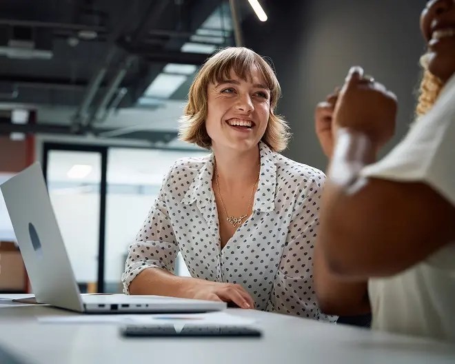 Smiling business professional with short hair and a white polka-dot blouse, talks with a coworker during a meeting, in a bright office.