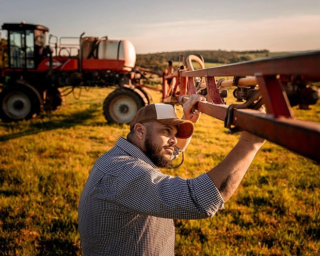 A man in a button-up shirt and cap inspecting farming machinery, with a tractor behind him.