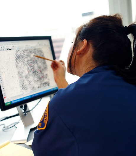 Female police officer in a blue uniform looks closely and points a pencil at a computer screen displaying a fingerprint.