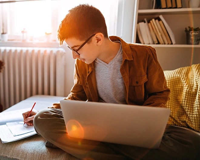 A student sitting cross-legged on a sofa with a laptop in his lap, writing in a notebook.