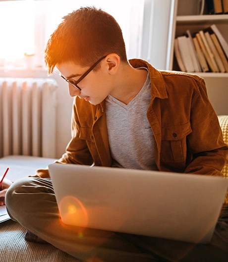 A student sitting cross-legged on a sofa with a laptop in his lap, writing in a notebook.