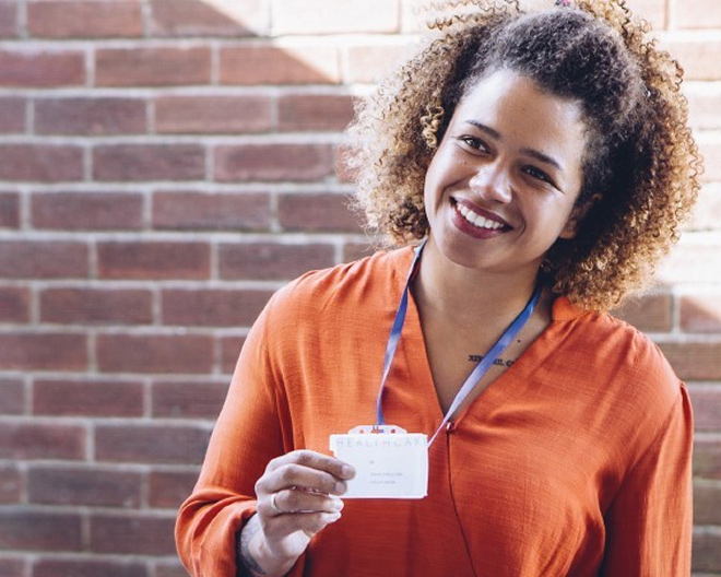 Smiling healthcare professional wearing an orange top, holding out her badge.