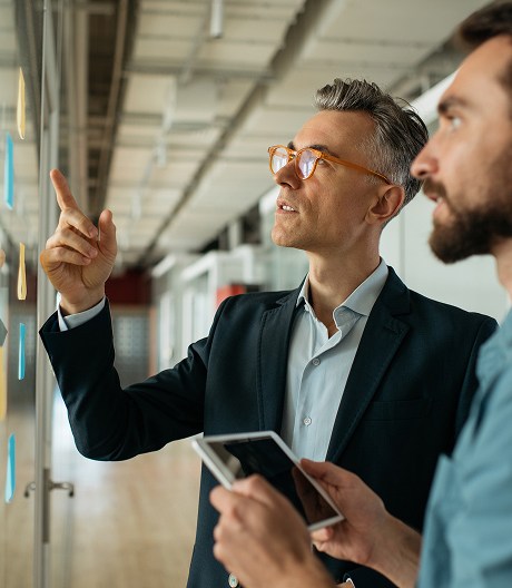 Male project manager in a suit and orange glasses points at colour-coded sticky notes on a glass wall while a colleague beside him holds a tablet.