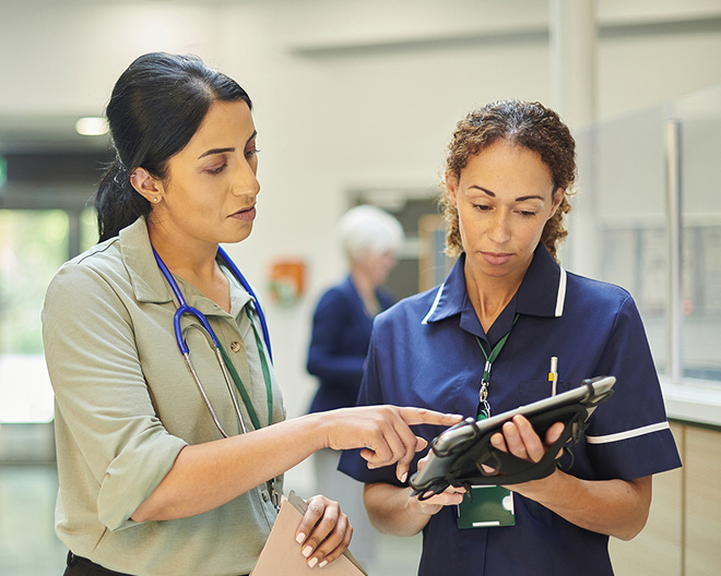 Young female doctor with a stethoscope and nurse look at information on a nurse’s tablet and discuss patient care in a hospital corridor.