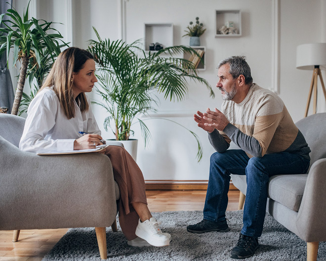 Female counsellor with notepad listens intently while a middle-aged man talks and gestures from a sofa in a calm, plant-filled office.