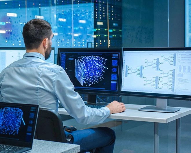 A man in a blue button-up shirt sitting at a desk with two monitors in a blue-lit office.