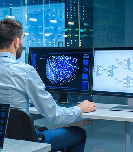 A man in a blue button-up shirt sitting at a desk with two monitors in a blue-lit office.