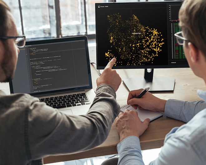 Close-up of a man’s hand pointing at a monitor displaying code, with a laptop beside it and another man looking on and taking notes.