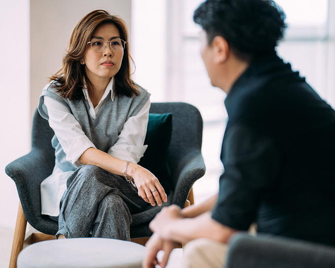 Professional woman in glasses sits with legs crossed, attentively listening to a male client during a one-to-one counselling session.