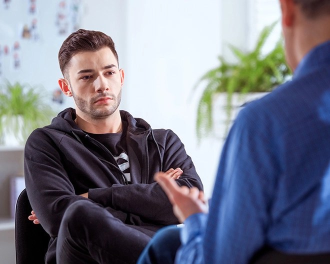 Young man sitting with arms crossed, listening to another man in a blue shirt who is speaking to him.