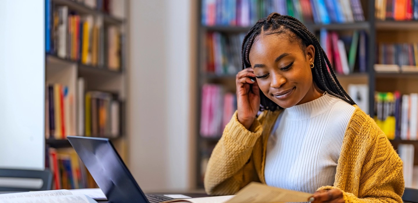 Smiling student in a mustard knit cardigan and white top pulls her hair back with one hand while reading a textbook in a bright, quiet library.