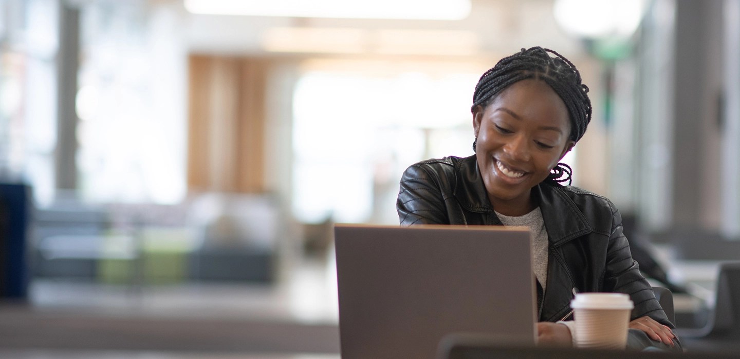 Smiling student in a leather jacket attentively taking notes during a lecture she's watching on her laptop.