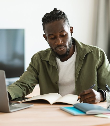 Young man studies at home, writing in an open notebook beside his laptop and textbooks on a tidy wooden desk.