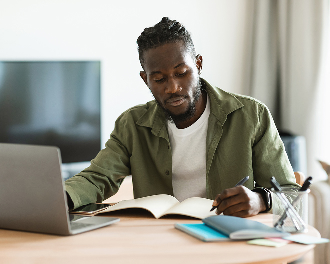 Young man studies at home, writing in an open notebook beside his laptop and textbooks on a tidy wooden desk.