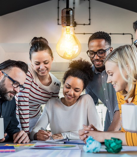Diverse group of colleagues crowd around a table, brainstorming and sketching ideas beneath an illuminated hanging lightbulb.