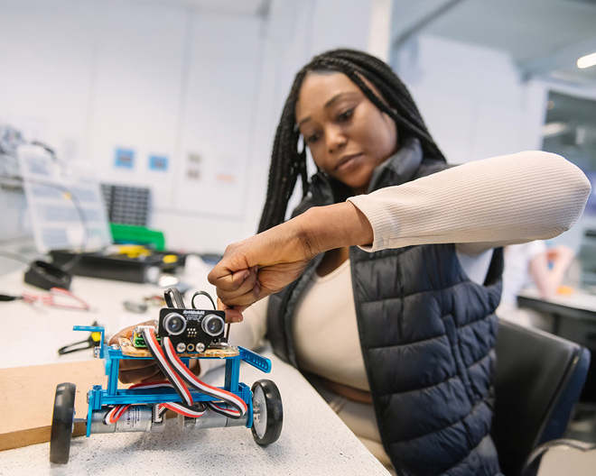 Female engineering student tightening a screw on a blue wheeled robot prototype in a university lab.