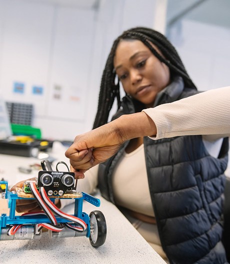 Female engineering student tightening a screw on a blue wheeled robot prototype in a university lab.