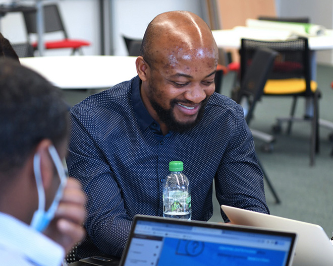 Smiling postgraduate reviews information on a laptop in a seminar room, water bottle beside him.