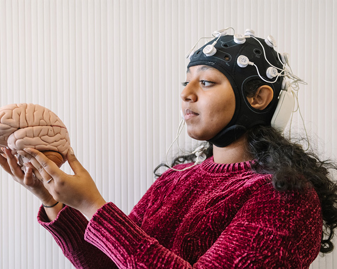 Young researcher wearing an EEG cap examines a plastic brain model against a neutral backdrop.