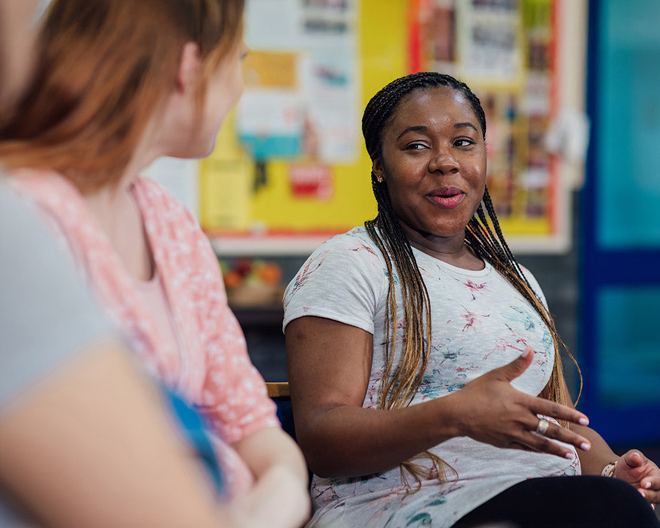 Woman sharing ideas during a small-group discussion on health behaviour change, gesturing while seated in a bright classroom.