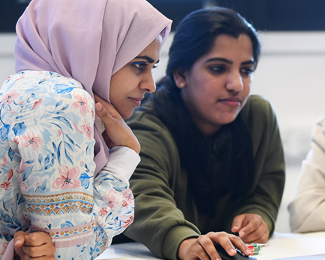 Two postgraduate students in conversation, one in a hijab, reviewing material on a computer for a health-promotion project.