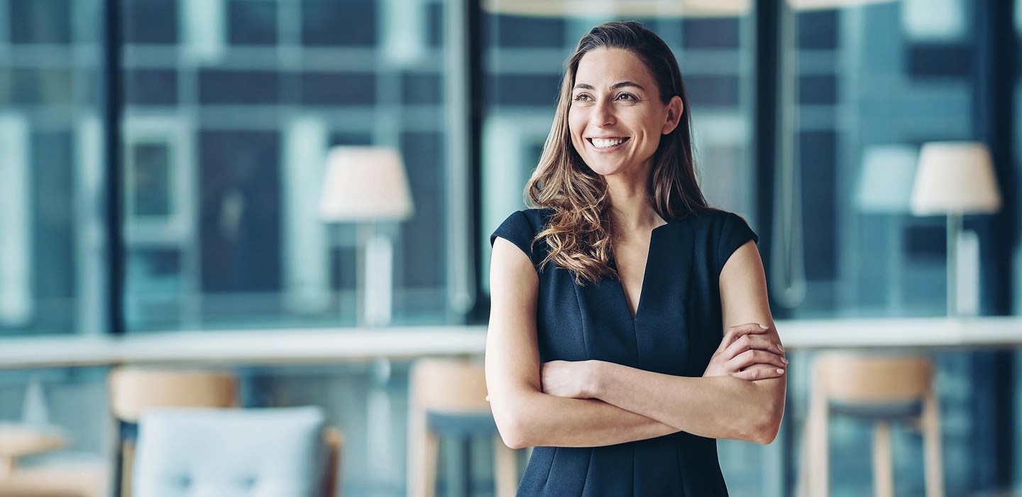 Close-up of a confident businesswoman in a business casual dress looks off to the side, smiling with crossed arms.