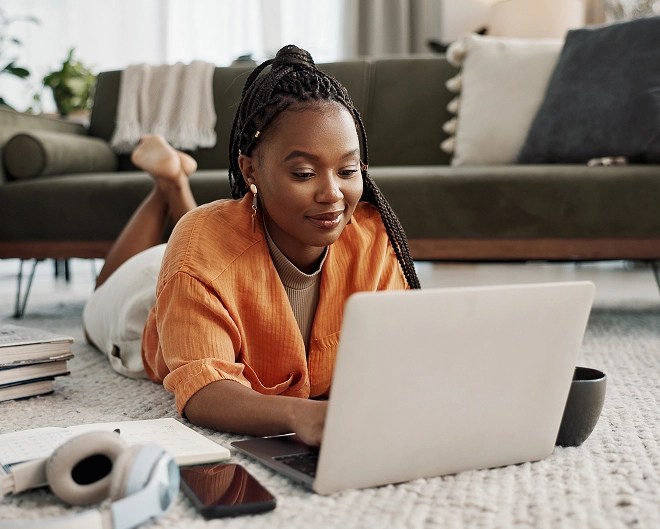 Online student wearing a bright orange cardigan, lying on the floor of a well-decorated apartment, working on her laptop with headphones, phone, and books nearby.