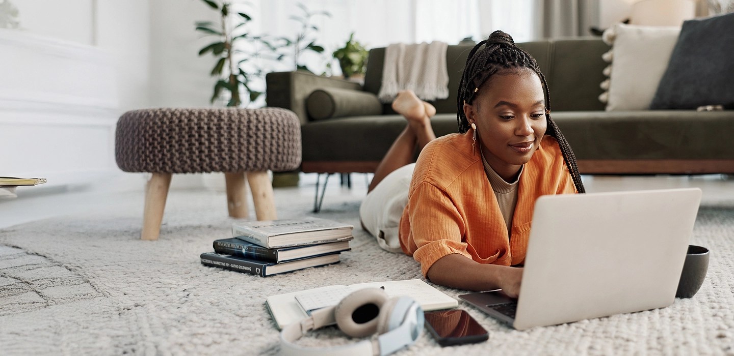 Online student wearing a bright orange cardigan, lying on the floor of a well-decorated apartment, working on her laptop with headphones, phone, and books nearby.