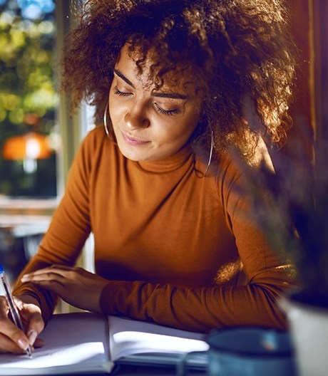 Young writer in a sunlit café thoughtfully drafting notes by hand in a journal.