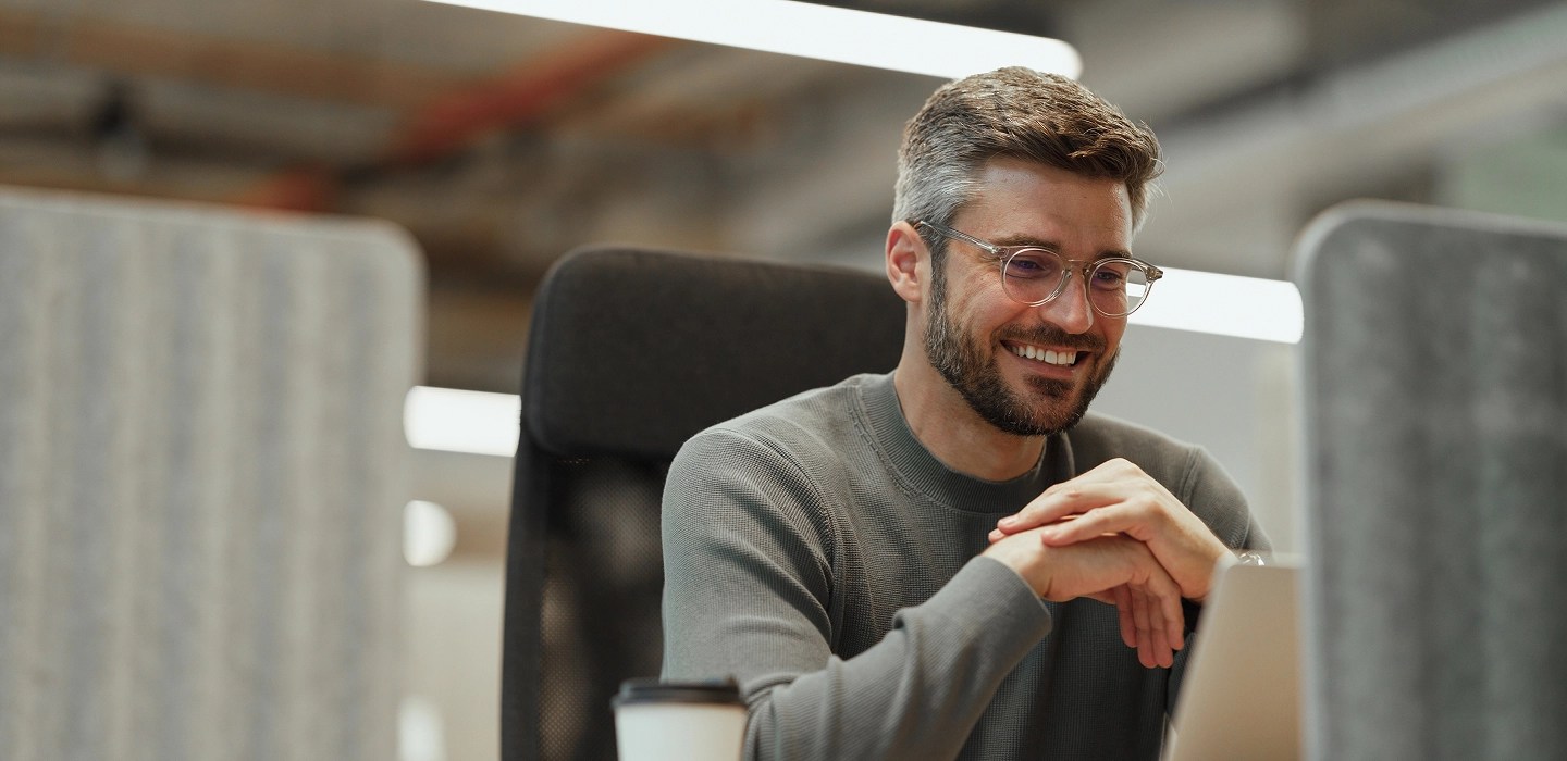 Casually-dressed professional with glasses smiles at his computer during a work meeting in bright office.