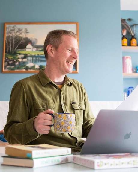 Man laughing at his laptop while holding a patterned mug and reading a script in a bright home kitchen.