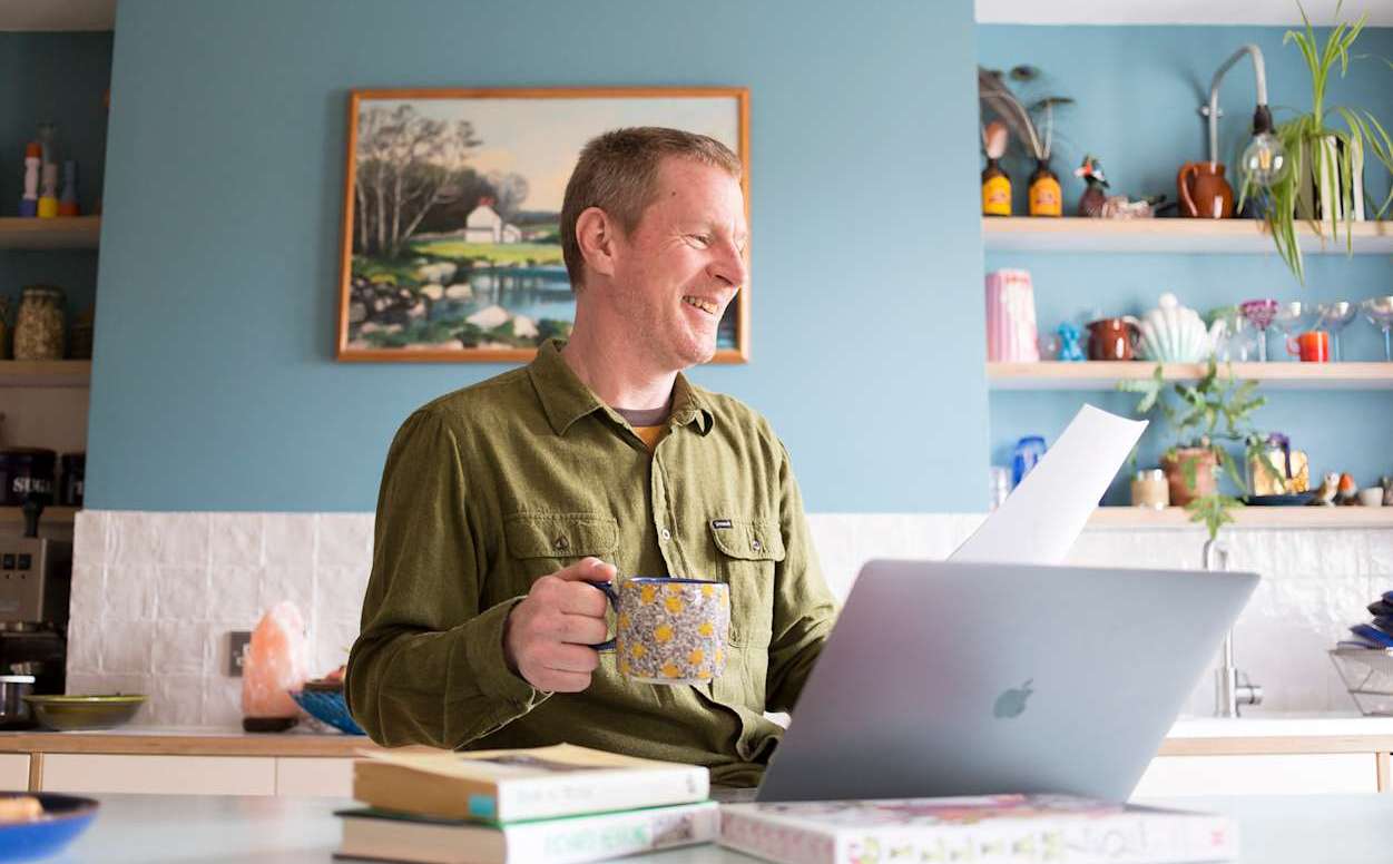 Man laughing at his laptop while holding a patterned mug and reading a script in a bright home kitchen.