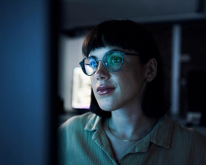 Close-up of an IT professional with short hair and glasses, illuminated by the glow of her screen as she concentrates in a dimly lit room.