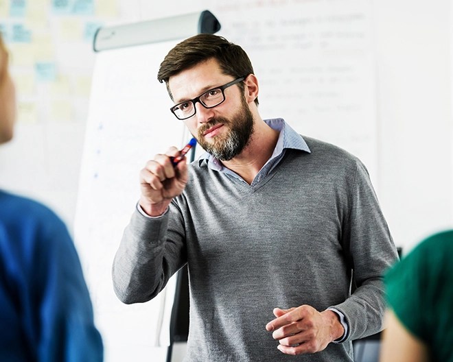 Bearded lecturer in glasses gestures with a pen while addressing students beside a flip-chart.