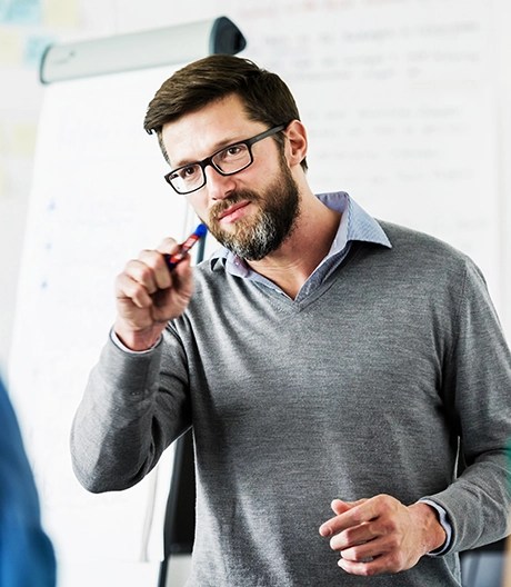 Bearded lecturer in glasses gestures with a pen while addressing students beside a flip-chart.