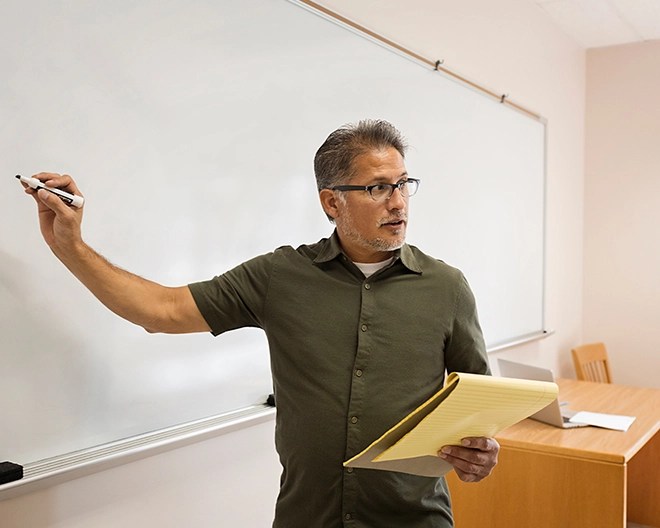 Mature teacher stands at a whiteboard, writing while holding a yellow legal pad during a classroom session.