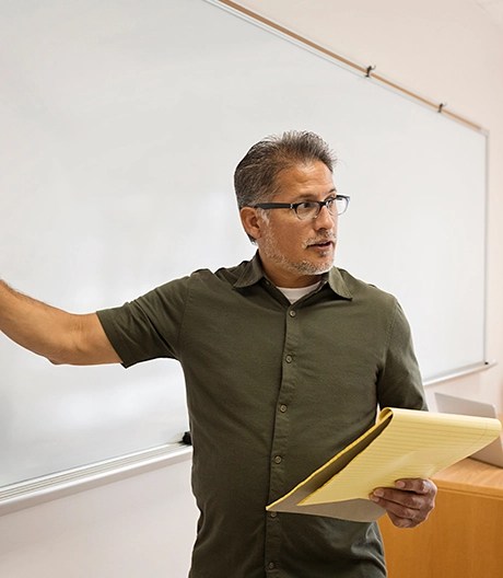 Mature teacher stands at a whiteboard, writing while holding a yellow legal pad during a classroom session.
