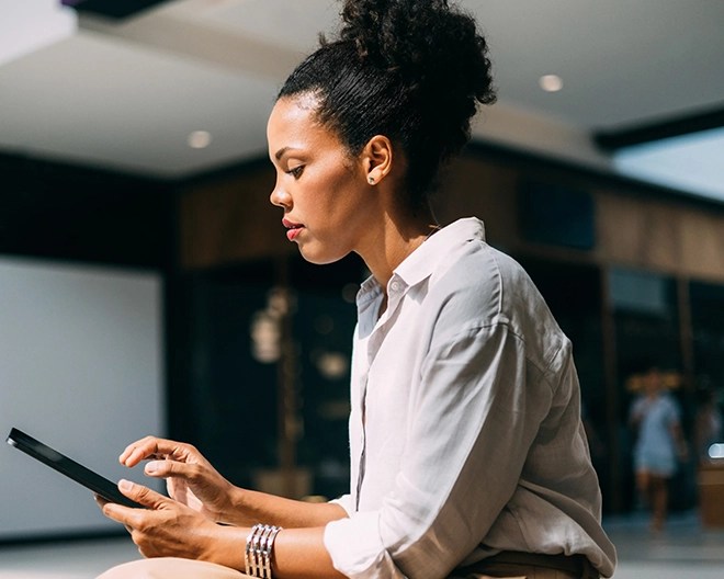 Focused young professional in a white blouse sits in a modern atrium, working on a tablet.