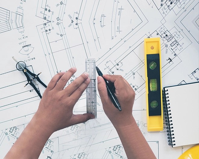 Close-up of hands drawing lines with a ruler on architectural blueprints, surrounded by drafting tools and a spirit level.