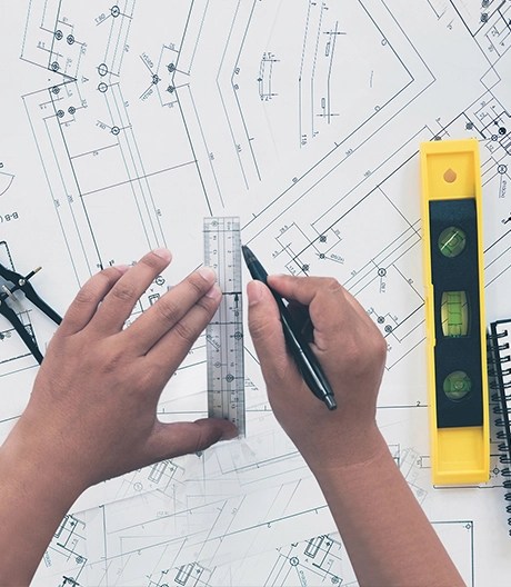 Close-up of hands drawing lines with a ruler on architectural blueprints, surrounded by drafting tools and a spirit level.