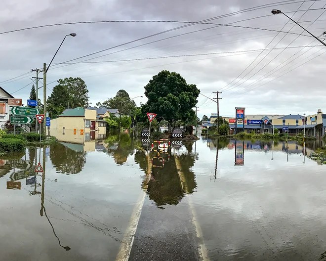 Small town main street completely flooded after heavy rain, with buildings, road signs and trees reflected in still water.