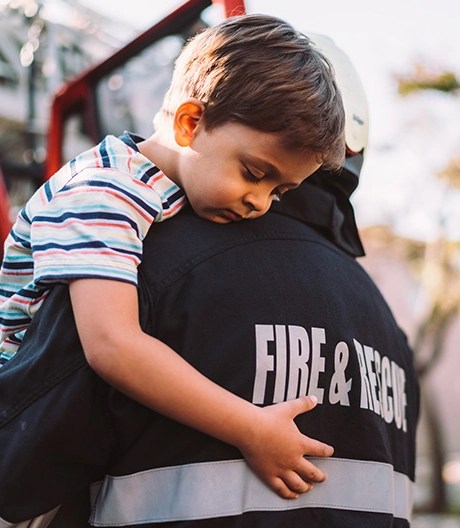 Fire & Rescue firefighter gently carries a young boy away from an emergency truck, the child resting his head on the responder’s shoulder.