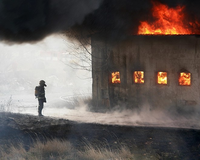 Smoke-shrouded firefighter stands before a warehouse engulfed in flames, bright firelight pouring from its windows.
