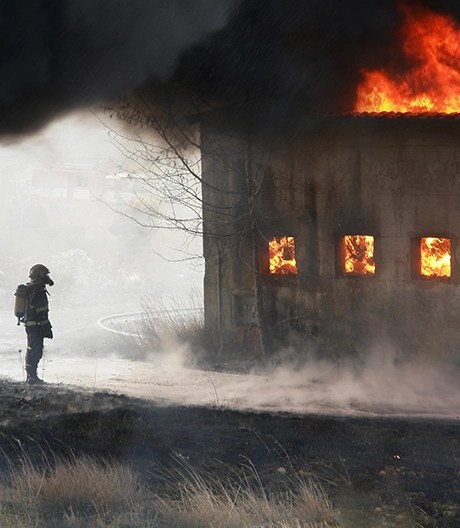 Smoke-shrouded firefighter stands before a warehouse engulfed in flames, bright firelight pouring from its windows.