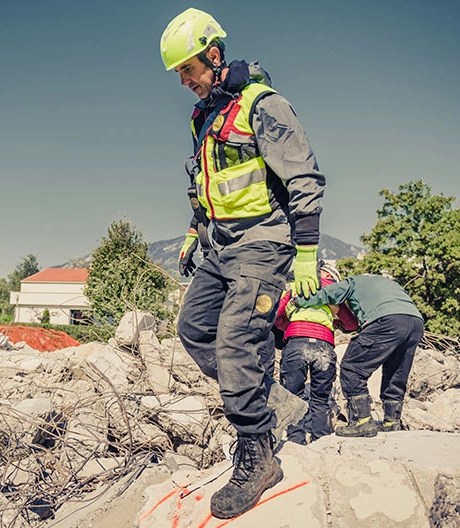 Emergency responder wearing a lime green helmet and vest walks through rubble, while colleagues assist a survivor in the background.