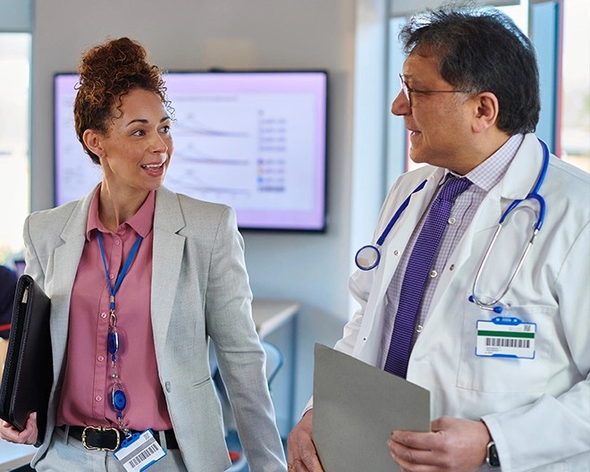 Doctor in a white coat with a stethoscope around his neck, holding a file, walking and talking to a healthcare manager in a blazer holding a portfolio inside an office.