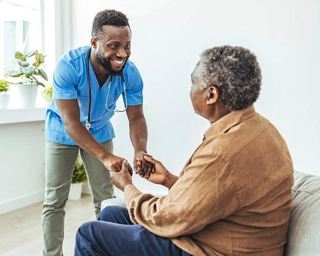Male nurse in blue scrubs smiling while holding the hands of an elderly patient.