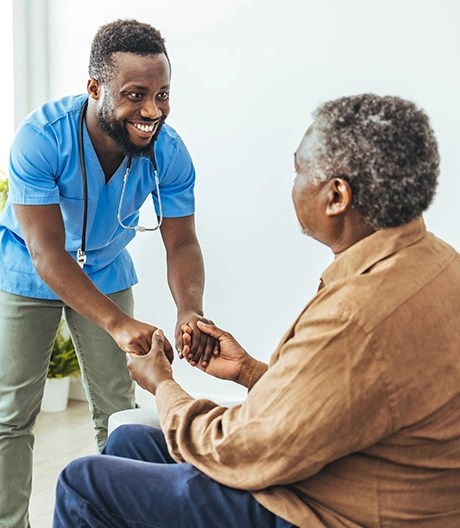 Male nurse in blue scrubs smiling while holding the hands of an elderly patient.