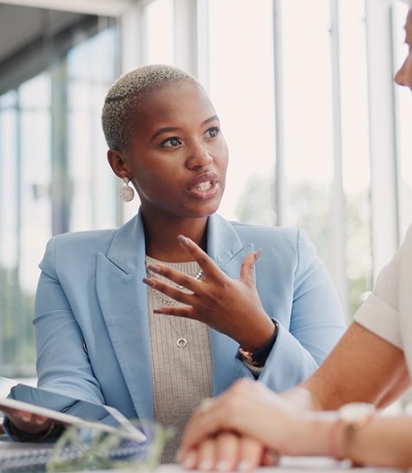 Marketing professional in a light-blue blazer explains ideas to a colleague, gesturing with one hand and holding a tablet.