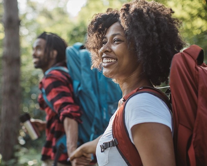 Smiling backpacker in a forest setting looks ahead while holding hands with a hiking companion, conveying sustainable travel.